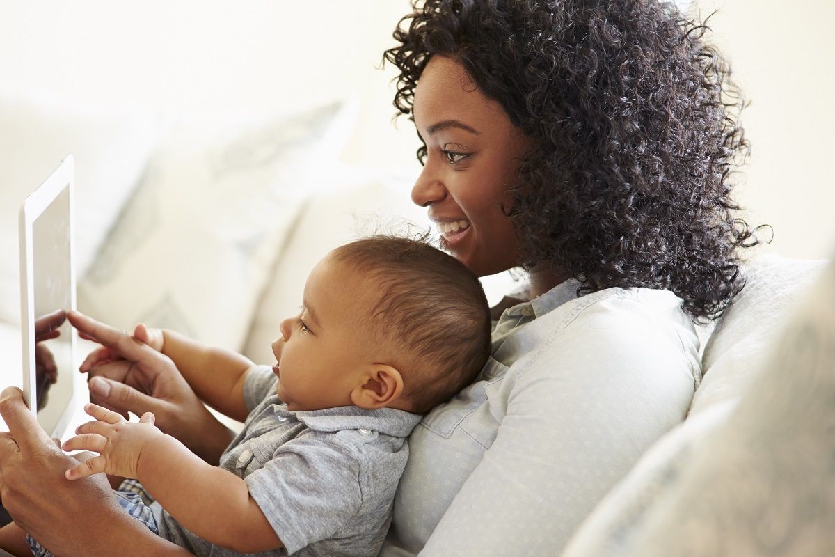 A woman with brown skin and curly hair holds a baby with brown skin. They both wear grey shirts and sit on a couch while looking at a tablet device.